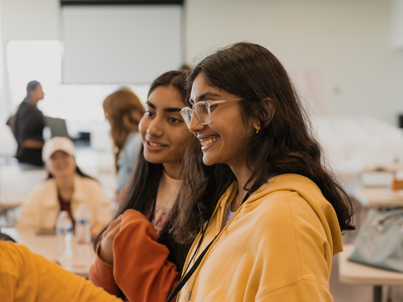 Two young women smile while other people converse in the background of a hackathon.